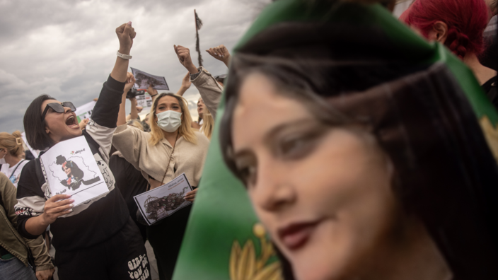 Im Hintergrund recken mehrere Frauen in einer Menschenmenge die Fäuste in die Luft und rufen. Sie halten Schilder in der Hand. Im Vordergrund ist eine Fahne mit dem Porträt der iranischen Kurdin Mahsa Amini zu sehen