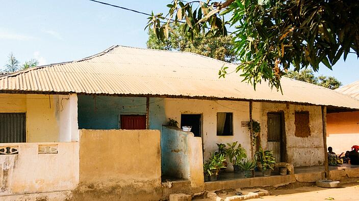A simple mud-plastered house with a corrugated iron roof stands slightly elevated in Guinea-Bissau, West Africa. The door is open. Potted plants stand in front of the door.
