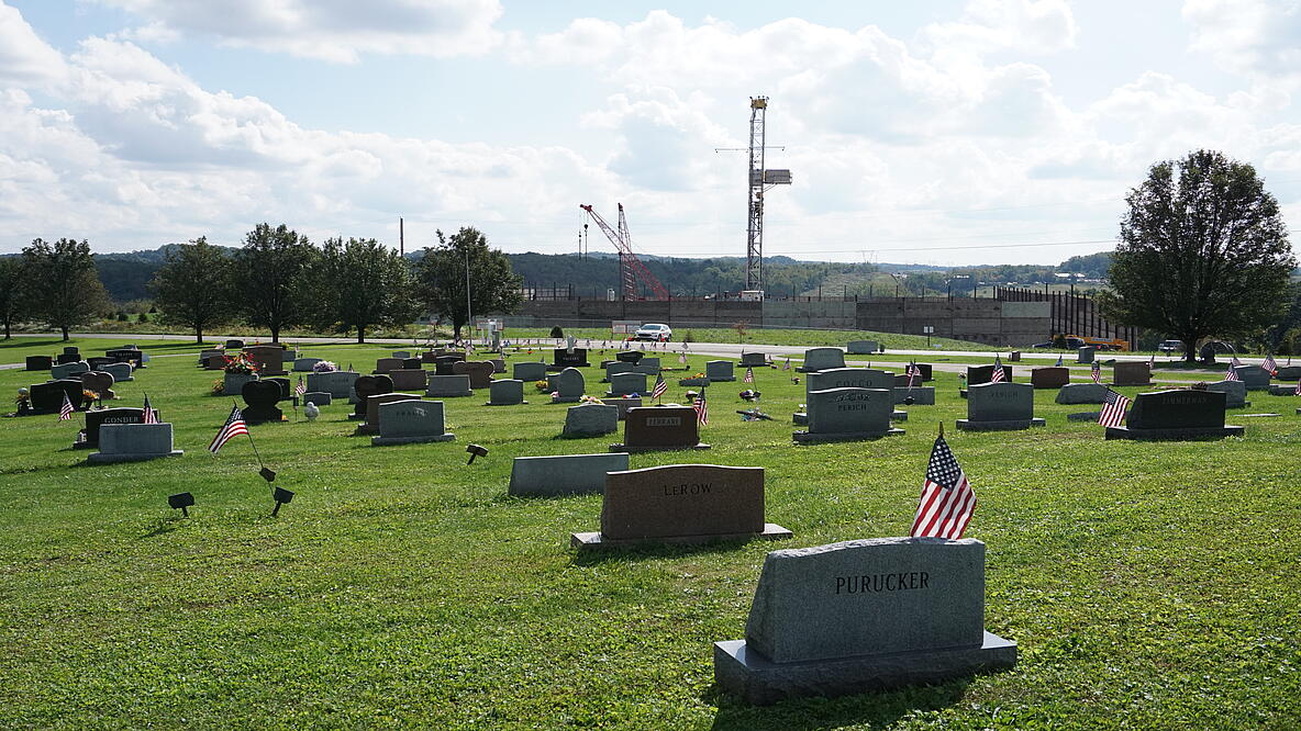 Ein Friedhof, die Gräber sind von hinten fotografiert. Viele Grabsteine sind mit der amerikanischen Flagge geschmückt. Hinter einer Absperrung stehen Bohrtürme und Kräne.