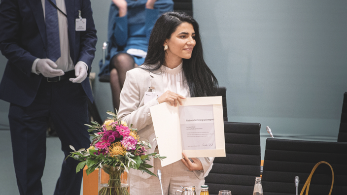 In an office environment, a young woman stands next to a bouquet of flowers and presents a certificate. She is wearing long black hair, a white trouser suit and a blouse. She looks to her right with a smile.