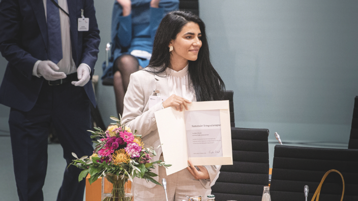 In einem Büroumfeld steht eine junge Frau neben einem Blumenstrauß und zeigt eine Urkunde. Sie trägt lange schwarze Haare, einen weißen Hosenanzug und eine Bluse. Sie blickt lächelnd zur rechten Seite hin.