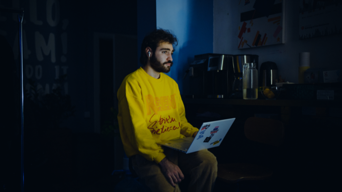 A man with a yellow sweater and a laptop on his knees sits in a dark room. He looks thoughtfully into the air, the scene is lit from the side by a pale light