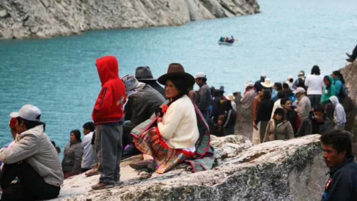 Peruvian women and men sit and stand on rocks at the edge of a blue lagoon.