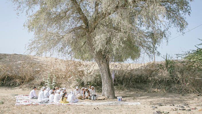 Ein sehr großer Baum steht inmitten einer kargen, sandigen Landschaft. Unter dem Baum sitzt eine Gruppe Inder auf ausgebreiteten Tüchern. Die meisten der Gruppe sitzen mit gesenktem Kopf.