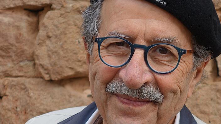 A color portrait of the writer Rafi Shami. He wears glasses, beret and smiles at the camera.