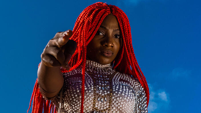 A young woman with long, red-colored braids poses in front of a deep blue sky with few clouds. She looks into the camera from above and points directly into it with two fingers