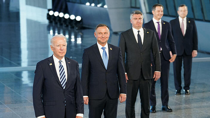 Five men in dark suits and white shirts stand diagonally behind each other with their arms hanging down. They smile and look past the camera on the left.