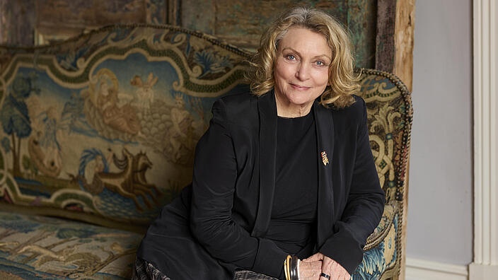 A slightly older lady sits on an opulent, carpeted sofa and rests her arm on the side rest. She looks friendly at the camera, her blonde mid-length hair is slightly wavy. She is wearing a black blazer with an insect-shaped brooch and many bangles. 