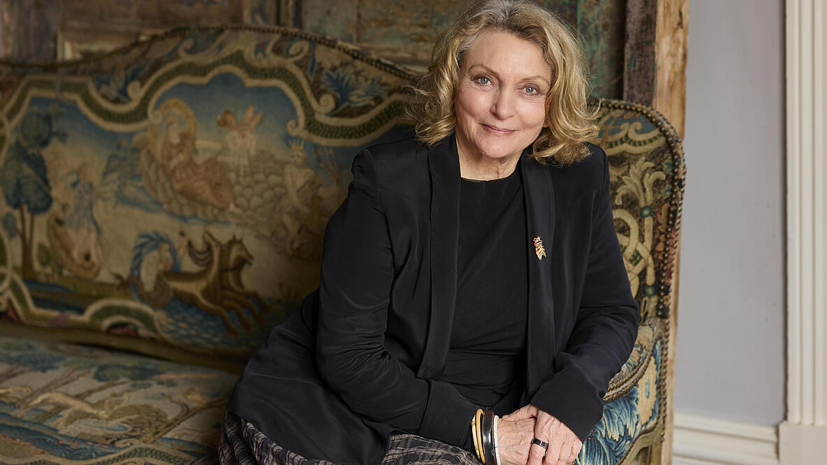 A slightly older lady sits on an opulent, carpeted sofa and rests her arm on the side rest. She looks friendly at the camera, her blonde mid-length hair is slightly wavy. She is wearing a black blazer with an insect-shaped brooch and many bangles. 