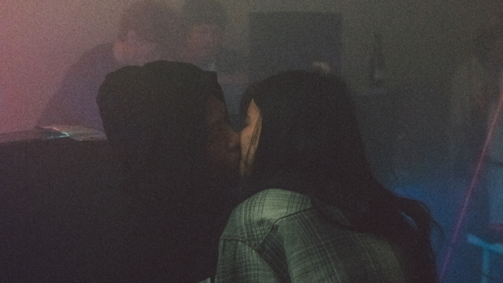 A club scene. In the background you can see two young people. In the front you can see a couple kissing. He is wearing a black hoodie. She has long black hair. It is dark.