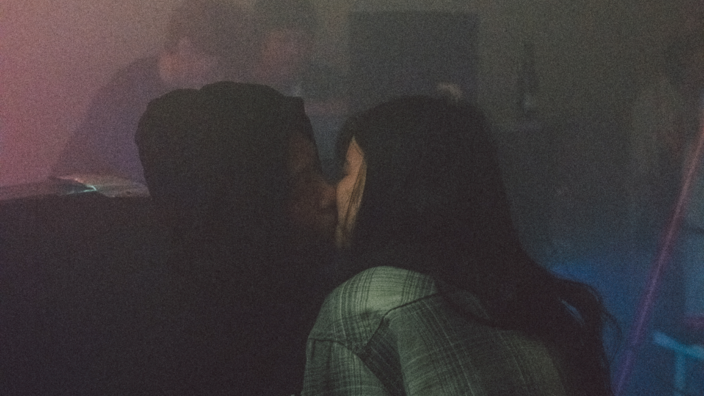 Eine Club Szene. Im Hintergrund sitzen zwei junge Menschen. Vorne sieht man ein sich küssendes Pärchen. Er trägt einen schwarzen Hoodie. Sie hat lange schwarze Haare. Es ist dunkel. 
