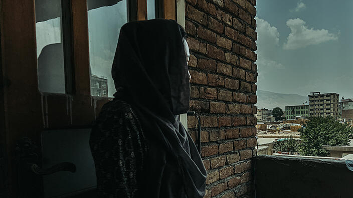 A woman is standing at the open window, her face turned away from us, looking into the distance. She wears a black headscarf and a patterned outer garment.
