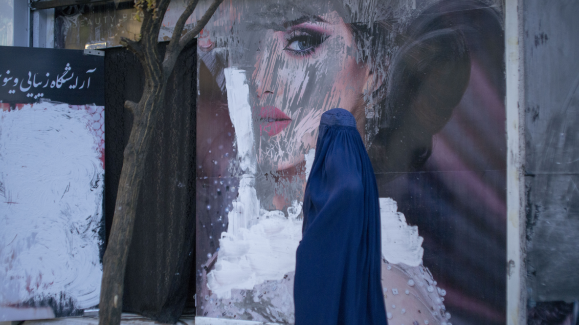 A woman in burqa walks past a billboard with outdoor advertising. On the poster is a very pretty made-up woman. The poster is smeared with white and gray paint.
