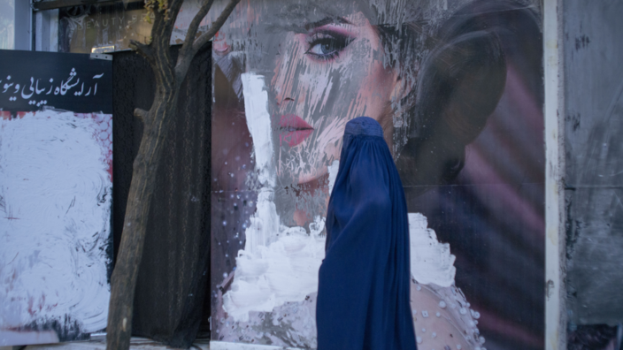 Eine Frau in Burka läuft an einem Plakat mit Außenwerbung vorbei. Auf dem Plakat ist eine sehr hübsche geschminkte Frau abgebildet. Das Plakat ist mit weißer und grauer Farbe beschmiert. 