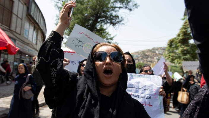 A woman is seen in front of a building wearing sunglasses and a black head scarf. She has raised her hand in protest and is chanting a slogan. She is surrounded by other protesters. 