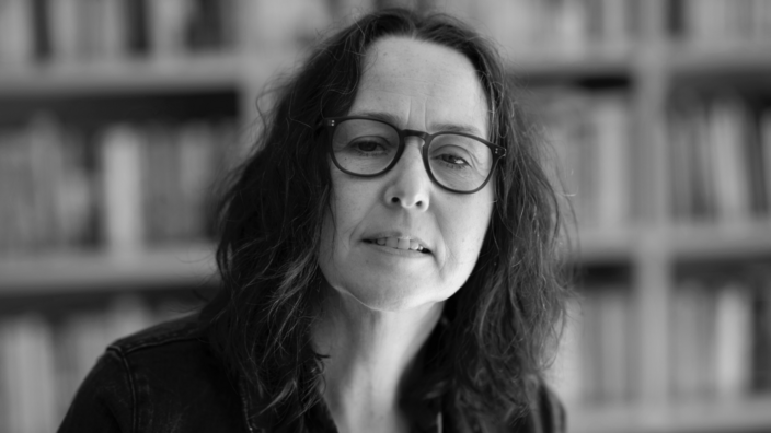 Black and white portrait of the author Audrey Magee in front of a bookshelf. She has long brown, slightly wavy hair and wears glasses.