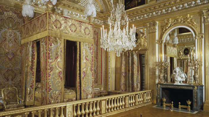 A golden bedroom, in the middle a richly decorated four-poster bed with red and gold curtains. On the right a fireplace, above it a mirror with a gilded frame, the walls are also covered with gold. From the high ceiling hangs a crystal chandelier.