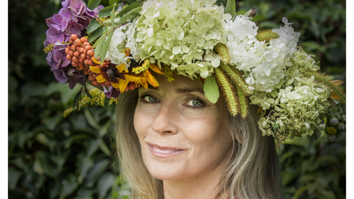 A side portrait of the writer Joanna Bator. She has long blonde hair. On her head she wears a large wreath of flowers and berries in white, green, orange and purple. She smiles at the camera.