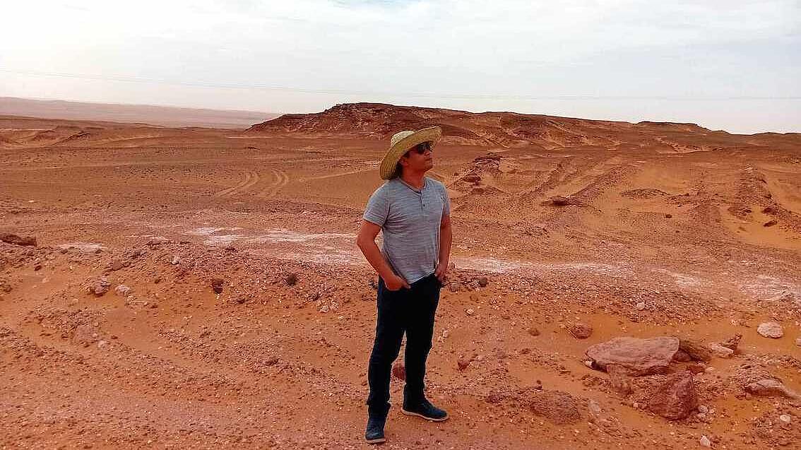 In einer Wüsten- und Gerölllandschaft steht ein Mann in Jeans und T-Shirt. Er trägt einen Strohhut mit breiter Krempe und Sonnenbrille, die Hände in den Hosentaschen und schaut in die Ferne. Die Landschaft ist leer, es ist keine Pflanze zu sehen  