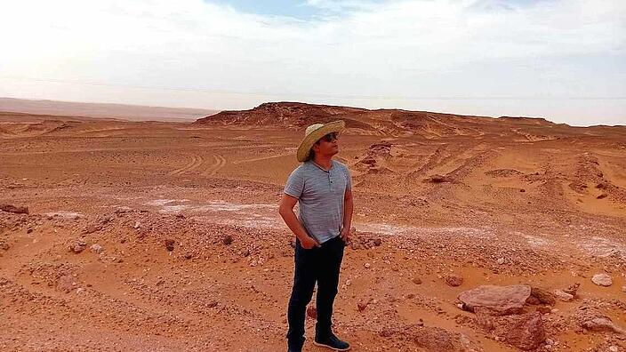 A man in jeans and a T-shirt is standing in a landscape of desert and grit. He is wearing a straw hat with a wide brim and sunglasses, his hands in his trouser pockets and looking into the distance. The landscape is empty, there is not a single plant to be seen