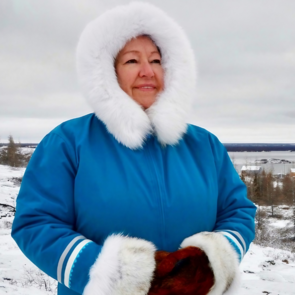 Eine Frau steht in einer winterlichen Landschaft. Sie trägt eine dicke Jacke mit Fellbesatz und dicken Handschuhen. Sie blickt in die Ferne, ihr Gesichtsausdruck ist selbstbewußt und heiter  