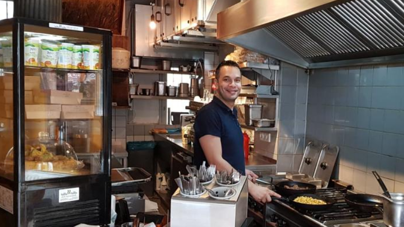 Ein Libanesischer Mann mittleren Alters steht am Tresen eines kleinen Restaurants. Er blickt freundlich in die Kamera