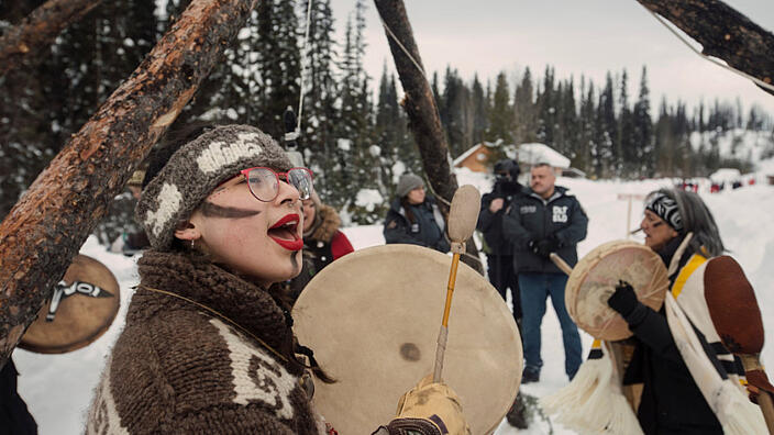 Drei Frauen stehen in einer Schneelandschaft umringt von Bäumen. Die Frauen singen und trommeln. Drei weitere Personen stehen am Rand und beobachten die Frauen.