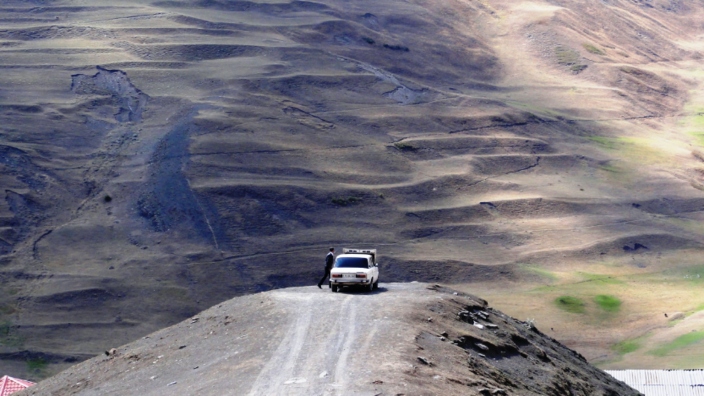 Ein einzelnes weißes Auto steht auf einem Berg vor einem Abgrund. Im Hintergrund erhebt sich ein Bergmassiv. 
