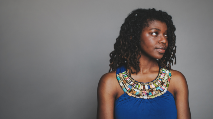 Ein seitliches Porträt einer jungen afrikanischen Frau mit gelockten Haaren. Sie trägt ein blaues Top mit bunter Perlenverzierung. 