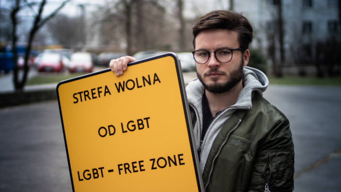 A young man with a full beard and glasses is standing on the street. He is holding a street sign up to the camera. The sign reads STREFA WOLNA, OD LGBT, LGBT - FREE ZONE.