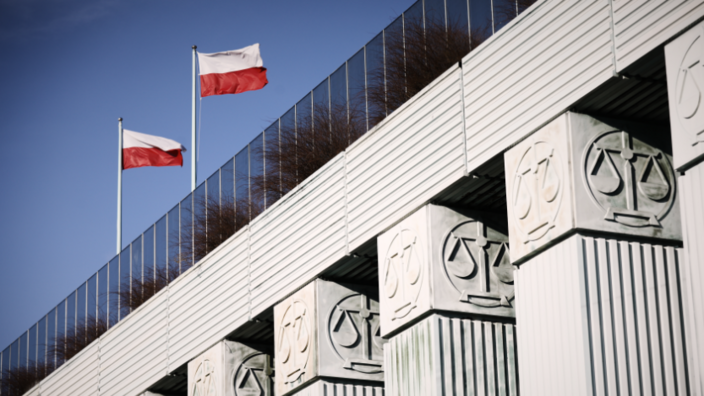 Ein Teil eines Gebäudes. Fünf rechteckige Betonsäulen, deren Abschluss am oberen Ende auf jeder Seite mit einer Waage verziert ist. Darüber die Verglasung des Gebäudes. Auf dem Dach wehen zwei polnische Flaggen in Rot und Weiß.