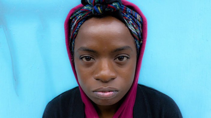 A portrait of a young black woman. Her gaze is fixed and a little defiant and indifferent at the same time. She wears a colorful scarf on her head, which is knotted over her forehead. Her hoodie in pink covers her colorful scarf at the back of her head. Over the hoodie in pink she wears another black top. The background is light blue. 