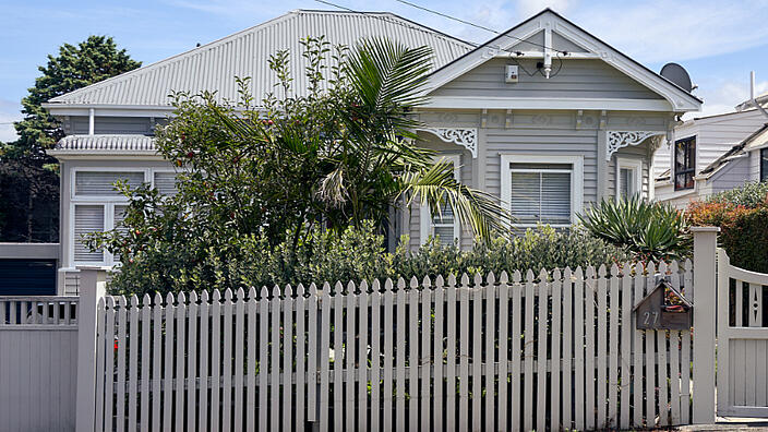 Behind a light gray garden fence stands a light gray house. The house is partially hidden behind bushes and a palm tree.
