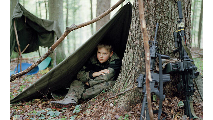 Ein etwa 14 jähriger Junge in Militär Uniform sitzt unter einer olivgrünen Plane an einem Baum. Er schaut unter der Plane hervor. Am Baumstamm lehnen zwei Waffen.
