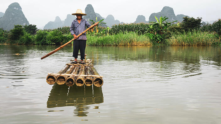 A medieval Chinese man wearing a straw hat stands on a raft made of six bamboo canes. The raft is on a body of water. At the edge of the water are palm trees and reeds. In the background you can see several mountains.