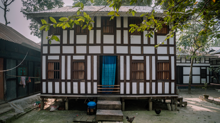 A white plastered house with two floors stands slightly elevated on stilts under trees in Bangladesh. A blue curtain is hanging in the central doorway.