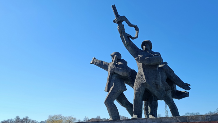 A monument of three men in Soviet military uniform in a victory pose stands against a blue sky. The foremost man holds his rifle in the air.