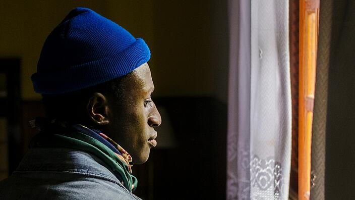 A side portrait of a young black man. He wears a denim jacket and a blue cap. He looks out of the window. The curtain is drawn to the side.