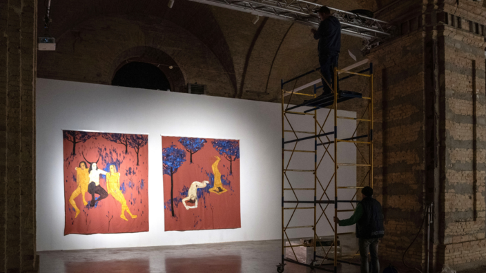 In a dark room with thick brick pillars there is an exhibition wall. Two paintings are hung on the wall, it is illuminated by a bright spotlight. In front of it some people are working, one of them is standing on a scaffold that reaches the ceiling