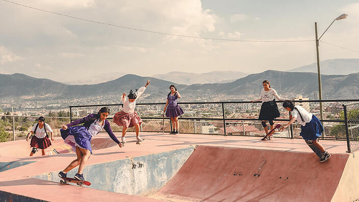 Junge Frauen skaten in traditionellen weiten, bunten Röcken in einem Skatepark in Bolivien. Der Platz liegt an einem Hang oben, so dass man einen sehr schönen Blick über ein bewohntes Tal hat. Der Himmel ist heiter bis wolkig