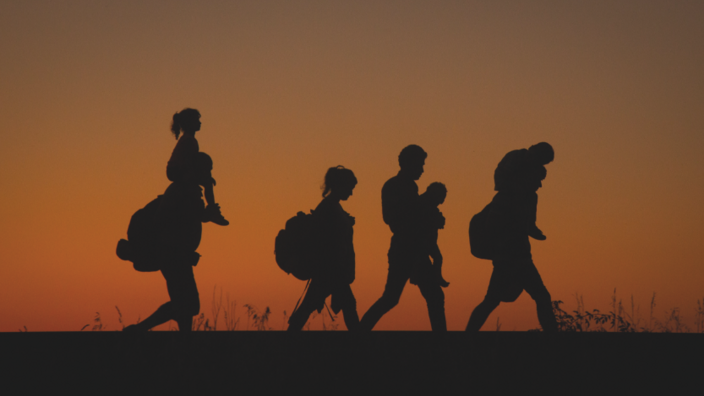 A group of people of different ages, packed with rucksacks, walk along a path. The group stands out against the orange-brown background like a play of shadows.