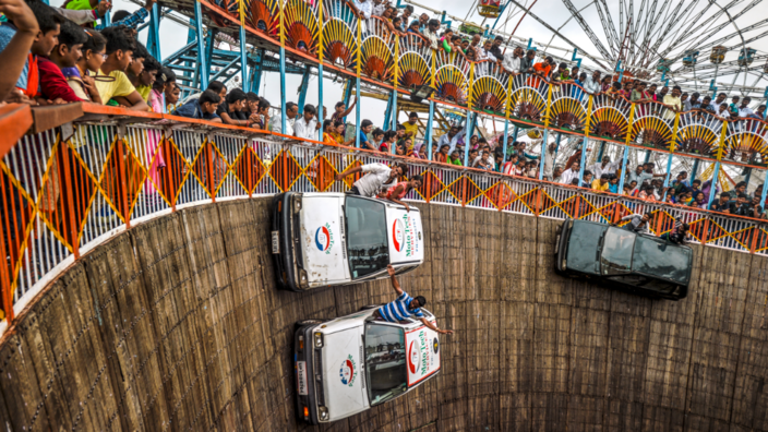 Three cars race along an almost vertical plank wall. The co-drivers hang out of the windows and cheer. Above the round are spectators in the stands. A Ferris wheel can be seen in the background.