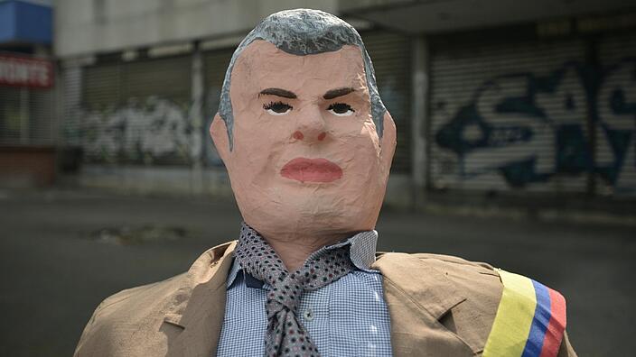 A human-sized papier-mâché doll that looks like Colombian President Ivan Duque. It is dressed in a jacket, shirt and tie. Over the left shoulder is a sash in the colors of the Colombian flag.