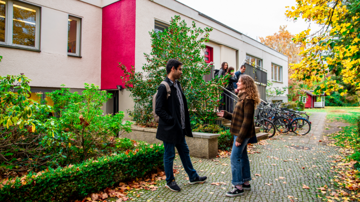 A male and female student are standing in front of a house and talking. In the background, three young people are standing on the steps in front of a house entrance. There are several bicycles at the foot of the stairs.