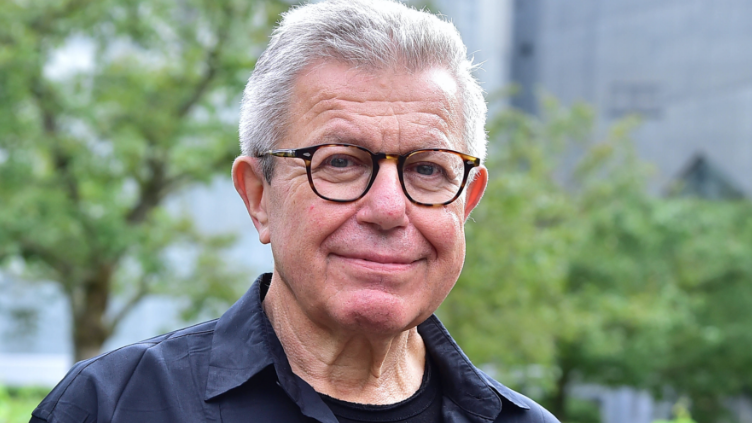 An older gentleman with glasses looks into the camera with a smile. Behind him trees and gray facades can be seen indistinctly. The gentleman wears white short hair, a black open shirt and a black t-shirt.