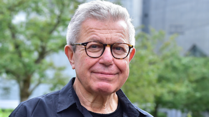 An older gentleman with glasses looks into the camera with a smile. Behind him trees and gray facades can be seen indistinctly. The gentleman wears white short hair, a black open shirt and a black t-shirt.