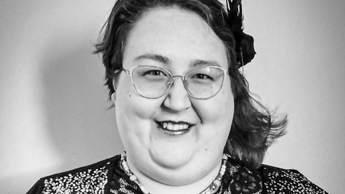 Ein schwarz weiß Fotoporträt von Bridget Liane. Sie trägt eine Brille, einen Federhaarreif und lächelt in die Kamera.