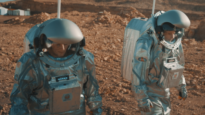 In a desert landscape two people in bulky silver space suits are walking around