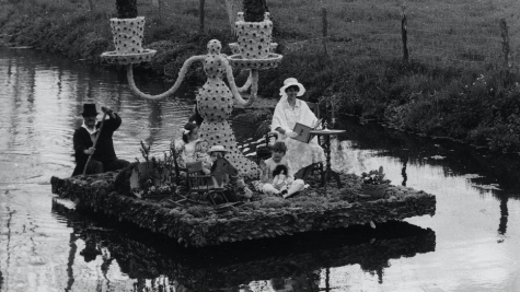 Ein Floß fährt auf einem Fluss. Eine Frau in weißer Kleidung sitzt auf dem Floß. Ein Mann mit Zylinder und schwarzem Mantel steuert das Floß. In der Mitte steht ein riesiger dreiflammiger Kerzenleuchter.