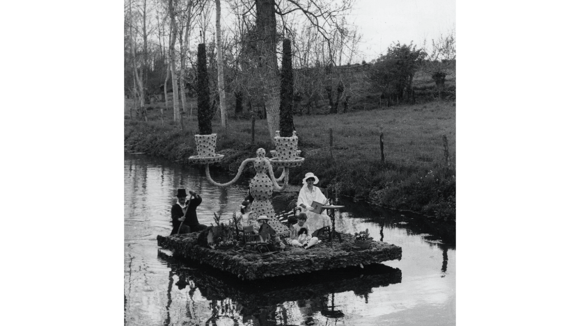 Ein Floß fährt auf einem Fluss. Eine Frau in weißer Kleidung sitzt auf dem Floß. Ein Mann mit Zylinder und schwarzem Mantel steuert das Floß. In der Mitte steht ein riesiger dreiflammiger Kerzenleuchter.